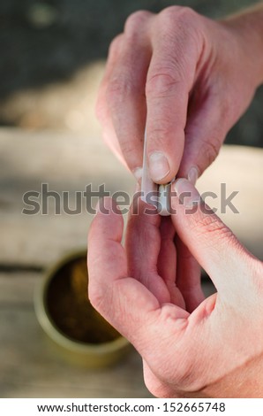 Man rolling himself a cigarette from loose tobacco blend using rollings, or adhesive paper covering sealed by licking, close up view of the hands