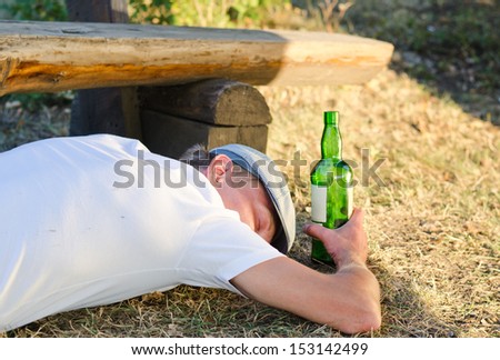 Drunk man holding a bottle of alcoholic beverage sleeping on the ground next to a bench in the park in summer