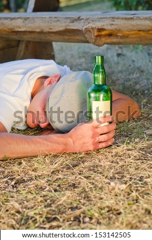 Drunk man holding a bottle of alcoholic beverage sleeping on the ground next to a bench in the park in summer