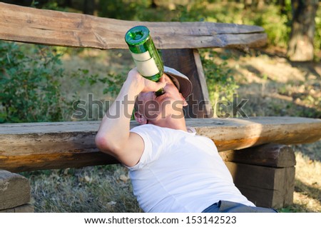 Jobless Caucasian alcoholic man drinking from a bottle of wine leaning on a bench in the park