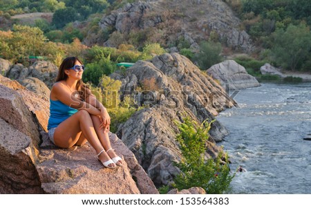 Attractive middle-aged woman relaxing in the mountains sitting on a rocky ledge overlooking a mountain river facing into the evening sun