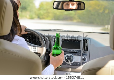 View inside a car from the back passenger seat of a woman driving holding a bottle of alcohol in one hand while steering
