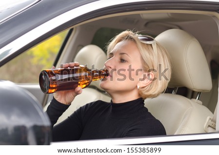 Young attractive blond female driver drinking alcohol from a bottle and driving the car with a smile of enjoyment on her face as she poses a threat to other road users