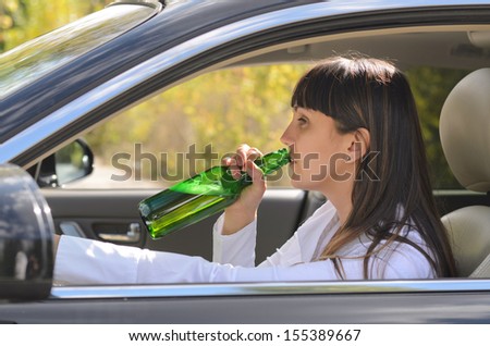 Alcoholic woman drinking and driving raising the bottle to her lips to take a swig as she steers the car, view through the side window