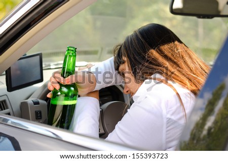 Drunk woman driver passed out in the car with her head resting on her arm on the steering wheel and her bottle of booze clasped in her hand
