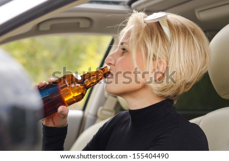 Young blond women driver drinking alcohol from a bottle and driving the car with a smile of enjoyment on her face as she poses a threat to other road users