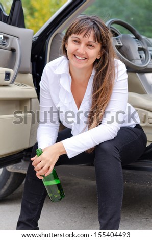 Happy woman drunkard in a car sitting on the sill of the open drivers door clasping her bottle of spirits and laughing