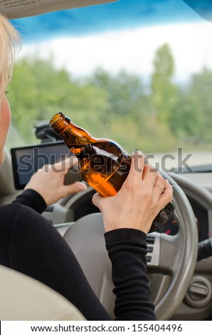 View inside a car from the back passenger seat of a woman driving holding a bottle of bear in one hand while steering