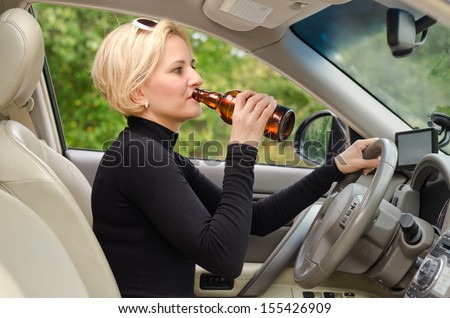 Young attractive blond female driver drinking alcohol from a bottle and driving sitting inside the car behind the steering wheel