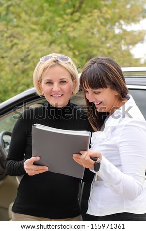 Two women signing a contract to buy a car standing in front of the vehicle with the paperwork and keys smiling happily