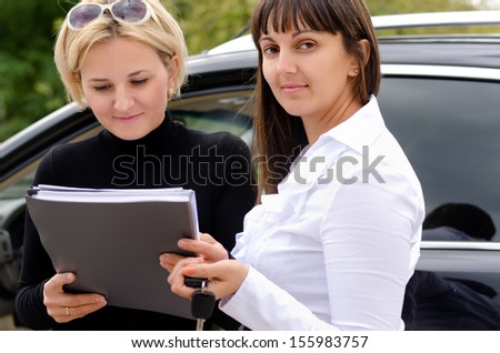 New attractive blond female owner purchasing a car from a smiling saleslady as they stand together alongside the vehicle signing the paperwork and handing over the keys
