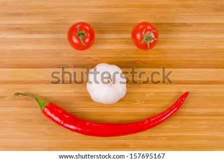 Vegetable face formed from a whole fresh red hot chilli pepper for a mouth, bulb of garlic for a nose and ripe red cherry tomatoes for eyes in a fun food background