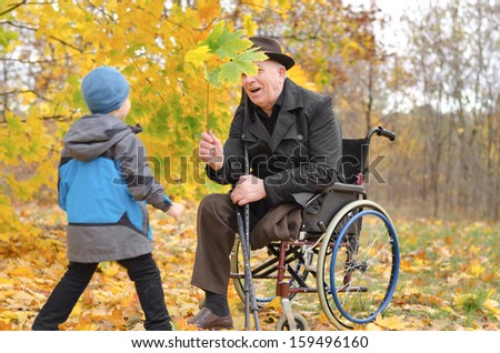 Young boy playing with his disabled grandfather as the two enjoy a day in the park together amongst the colourful autumn leaves running towards his wheelchair