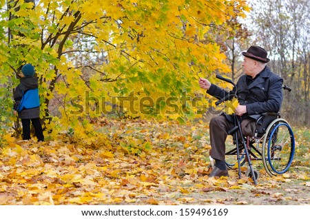 Disabled grandfather using wheelchair taking care of his playing grandchild outdoors in the forest in the autumn
