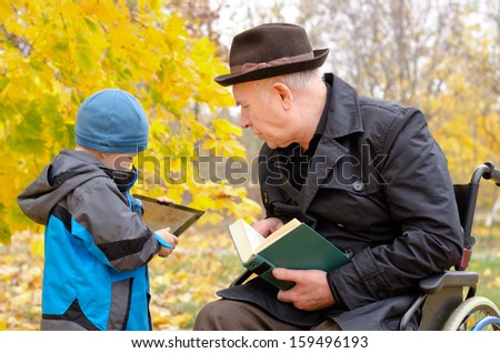 Generation gap between grandchild using a modern tablet pc and disabled grandfather in a wheelchair holding a book