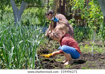 Cute little boy weeding the vegetable garden crouching down alongside his mother in the summer sunshine with his plastic spade
