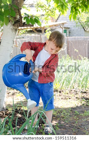 Little boy watering the vegetables standing in the shade of a tree in the veggie garden on a hot summer day pouring from a large blue plastic watering can