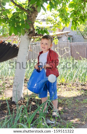 Cute little boy helping out in the vegetable garden standing in the shade of a tree holding a large blue plastic watering can and smiling happily
