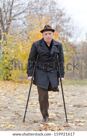 Disabled senior man on crutches determined to continue to enjoy a healthy active lifestyle enjoying a walk in an autumn park in his coat and hat