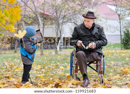Disabled grandfather sitting in a wheelchair watching his young grandson playing with a tablet computer as the two enjoy a relaxing day in an autumn park together