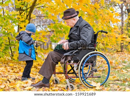 Elderly disabled man sitting reading a book in a wheelchair enjoying a day out in an autumn park with his little grandson who is standing playing with a tablet computer