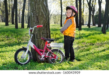 Little boy standing grinning alongside his bicycle which is propped against a tree in a wooded park as he looks over his shoulder at the camera