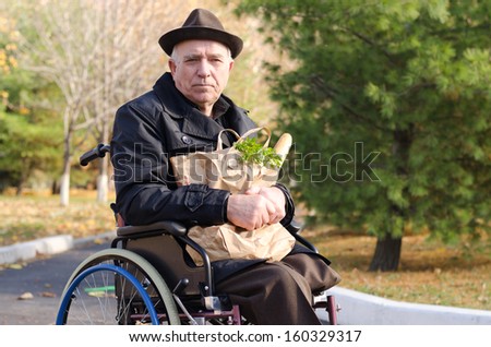 Senior handicapped man doing his grocery shopping sitting in the road in his wheelchair clutching a large bag of food and looking at the camera