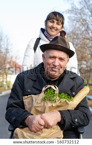 Smiling senior man sitting in a wheelchair with a bag of groceries on his lap being pushed along the street by a daughter or carer