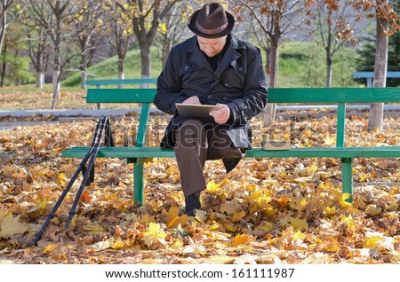 Elderly handicapped man on crutches sitting on a wooden bench dressed in a warm overcoat against the chilly autumn weather using a tablet computer in the park