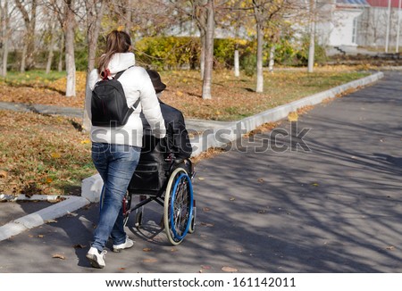 Female carer taking a disabled man in a wheelchair for a walk along the street as they walk away from the camera