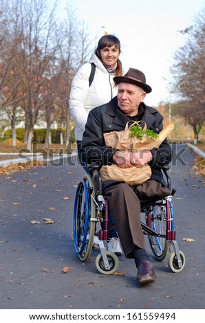 Smiling senior man sitting in a wheelchair with a bag of groceries on his lap being pushed along the street by a daughter or carer