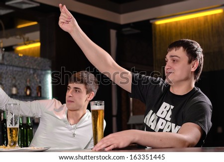 Two attractive young men sitting at the counter drinking a pint of beer calling for attention in the pub with their hands raised in the air