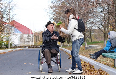 Mother helping her elderly disabled father in his wheelchair with his grocery shopping as her young son sits on the wooden park bench watching the two of them