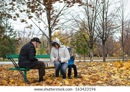 Disabled elderly man with crutches and an younger woman playing chess sitting together on a wooden park bench wrapped up warmly against cold autumn weather