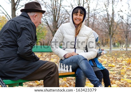 Attractive smiling woman sitting on a park bench playing chess with an elderly man while her young son amuses himself on a tablet computer behind her