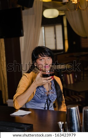 Woman drinking alone at the bar sitting at the counter holding a large glass of red wine in her hand as she looks at the camera with a thoughtful expression