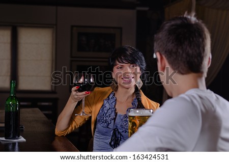 Smiling young woman chatting to her boyfriend at the bar leaning on the counter with a large glass of red wine in her hand as she smiles at him