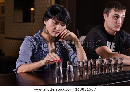 Lonely woman alcoholic sitting at a bar counter with a long line of full shot glasses staring morosely at the counter as she drinks her way through them