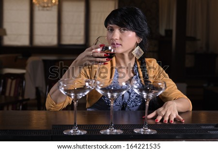 Lonely stylish young woman sitting drinking alone at a bar in a hotel or nightclub, view with three cocktail glasses in the foreground