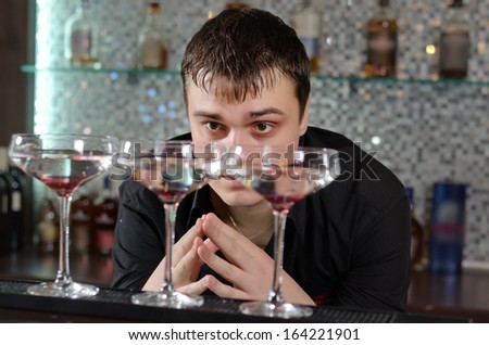 Attractive young man bending down and checking out three glasses of cocktail standing on a bar counter at a hotel or nightclub