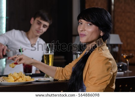 Young woman enjoying a snack at bar sitting eating chips close by as the bartender works preparing drinks in the background