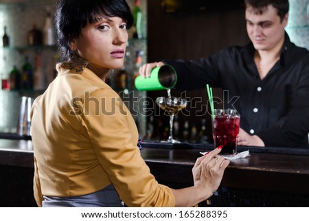 Woman sitting at a bar waiting to be served looking back over her shoulder at the camera as the barman mixes her a cocktail