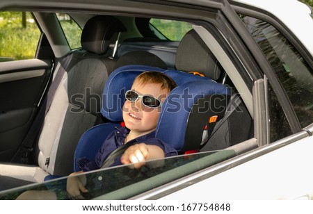Little boy in trendy sunglasses relaxing in the back of a car in a child safety seat as he waits for his parents , view through the back passenger window