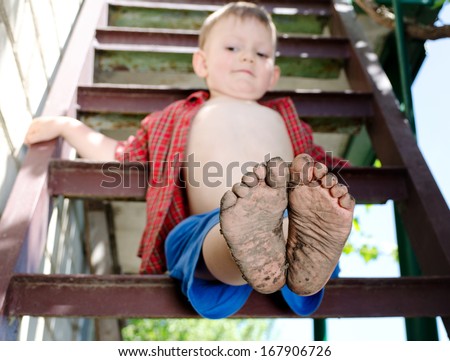 Little boy showing off his dirty feet sitting on a step holding them out to the camera with their muddy soles after playing barefoot in the garden