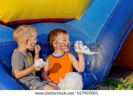 Two young boys sitting close together on a plastic jumping castle at a funfair eating candy floss together smiling at the stickiness of the spun sugar