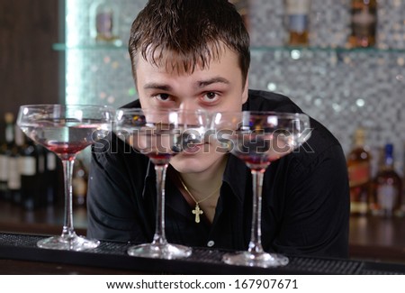 Barman working behind the bar counter peering across at the camera between three cocktail glasses lined up on the counter