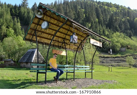 Man sitting on a covered bench in a field with an attached clock and loudspeaker against a backdrop of forested mountains and a timber cabin