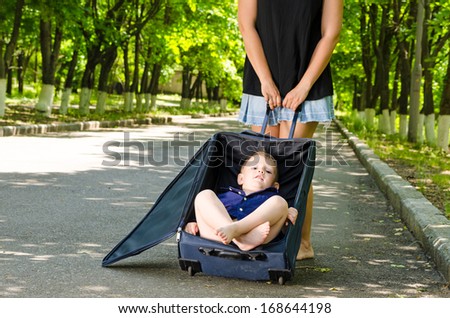 Little boy waiting with his mother in a country road sitting in an open suitcase on the asphalt