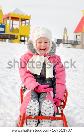 Pretty little girl dressed in a fashionable warm pink winter outfit playing on a sled in the snow reclining on her back smiling happily at the camera