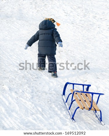 Little boy pulling a blue sled behind him on the snow as he trudges up the slope for a new descent on a cold winter day
